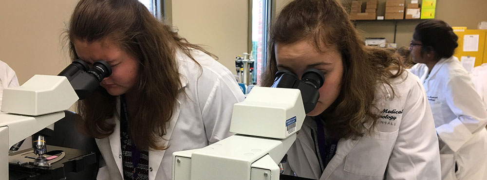 Students in white coats peering into microscopes in a lab.