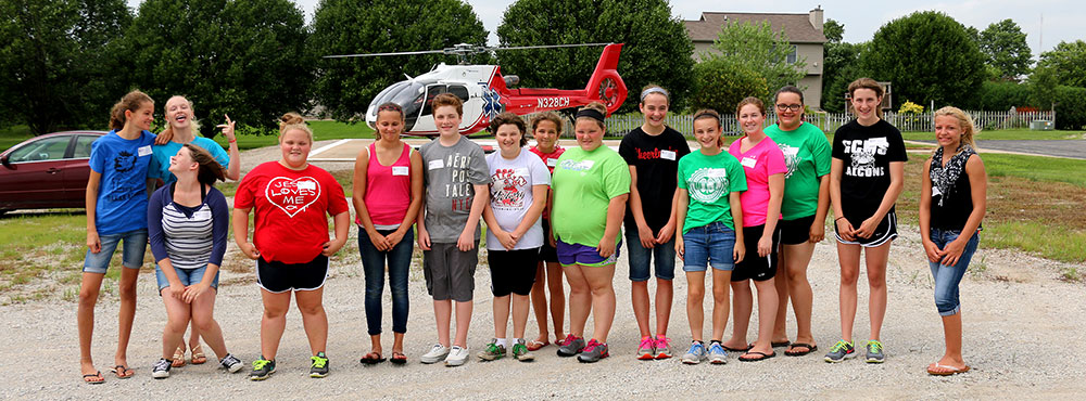 Kids in shorts and T-shirts pose in front of a helicopter.