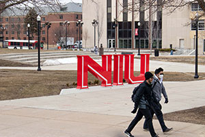 Two warmly dressed students walk in front of the large metal NIU logo on campus.