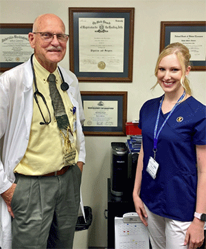 Participant and older medical professional pose in front of his wall of diplomas.